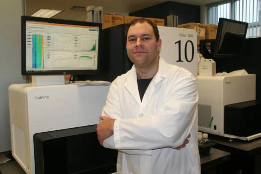 Yaron in a lab coat beside a DNA sequencer at the Genome Sciences Centre